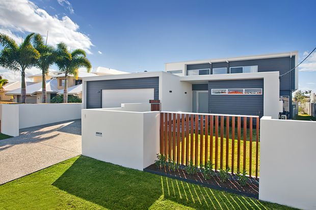 Contemporary two-storey suburban house exterior with a white rendered facade, dark grey horizontal cladding, integrated garage, flat rooflines, and broad horizontal windows. A timber slat front gate and low rendered boundary walls frame the landscaped lawn and entry path, with palm trees and a bright blue sky in the surroundings.