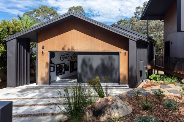 Contemporary detached garage or outbuilding with a prominent pitched gable roof, dark metal framing and cladding, and a warm timber-lined front facade. The wide opening reveals bicycle storage and framed items inside. A landscaped forecourt with exposed rocks, ornamental grasses, stepping stones, and planting connects it to a neighbouring dark-clad structure.
