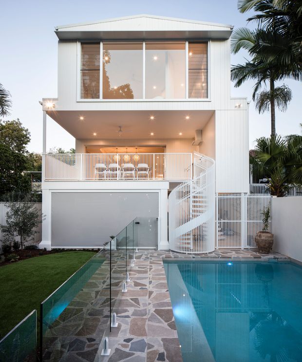 Exterior view of a contemporary multi-storey residence with white vertical cladding, a broad upper-level glazed window, an open entertaining balcony, and a distinctive white spiral staircase. A swimming pool, irregular stone-paved path, glass pool fencing, and tropical planting occupy the foreground.