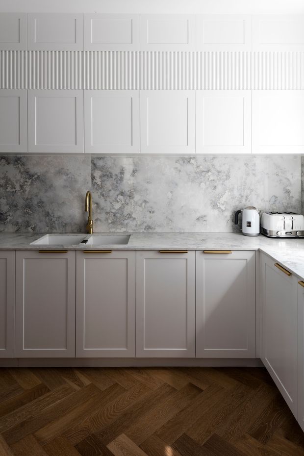 A refined kitchen with pale grey shaker-style cabinetry, brass handles and tapware, a dramatic cloudy grey stone backsplash and benchtop, white upper cabinets with decorative fluted detailing, integrated sink, small appliances, and dark timber herringbone flooring. Natural light creates a soft, elegant atmosphere.