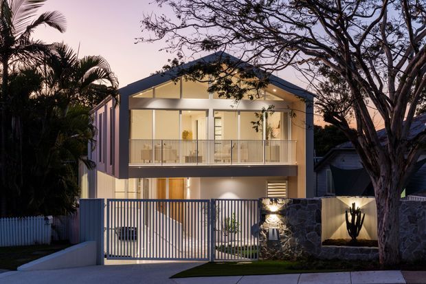 Twilight exterior view of a contemporary two-storey residence with a prominent gabled roof, illuminated upper-level balcony, white rendered facade, vertical metal front gate, and low stone boundary wall. Tropical planting and mature trees frame the suburban frontage.