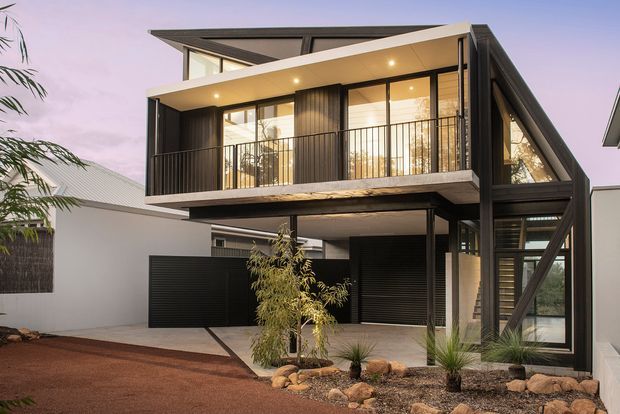 Exterior view of a contemporary multi-storey residence at dusk, featuring a bold angled roof and projecting upper-level balcony. The facade combines dark vertical cladding, extensive floor-to-ceiling glazing, black metal balcony balustrades, and a ground-level garage or service frontage. A landscaped forecourt with gravel, rocks, and drought-tolerant planting sits in front of the home.