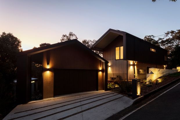Twilight exterior view of a contemporary multi-level residence on a sloping suburban site. The home features dark vertical cladding, contrasting warm timber-toned illuminated walls, steep intersecting pitched roof forms, a prominent garage volume, and a sloped concrete driveway with integrated landscape planting and exterior lighting.