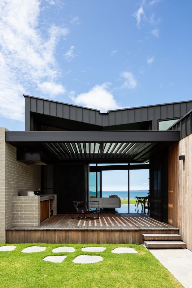 A modern coastal home exterior viewed from a small lawn, featuring a dark metal-clad roof and upper facade, exposed structural beams, a covered timber deck, and full-height glazed doors opening to a compact living area. A built-in brick outdoor kitchen or barbecue sits to the left, while the ocean is visible beyond the glazing. The design combines strong geometric lines, timber cladding, black metal framing, and a slatted pergola roof.
