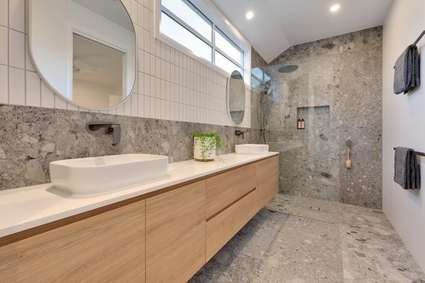 Contemporary bathroom with a long floating timber vanity, two white countertop basins, round mirrors, and brushed metal wall-mounted tapware. Large-format grey stone tiles cover the floor and shower walls, with a frameless glass shower enclosure and high clerestory windows providing abundant natural light. Dark towels are mounted on the white side wall.