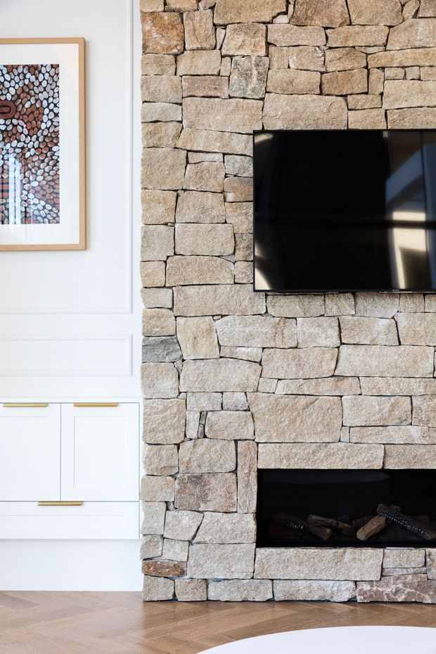 Close-up interior view of a living room feature wall combining a large rough-cut natural stone fireplace surround with a black wall-mounted television. White paneled cabinetry with brass pulls, framed artwork, and herringbone timber flooring are visible alongside the fireplace.