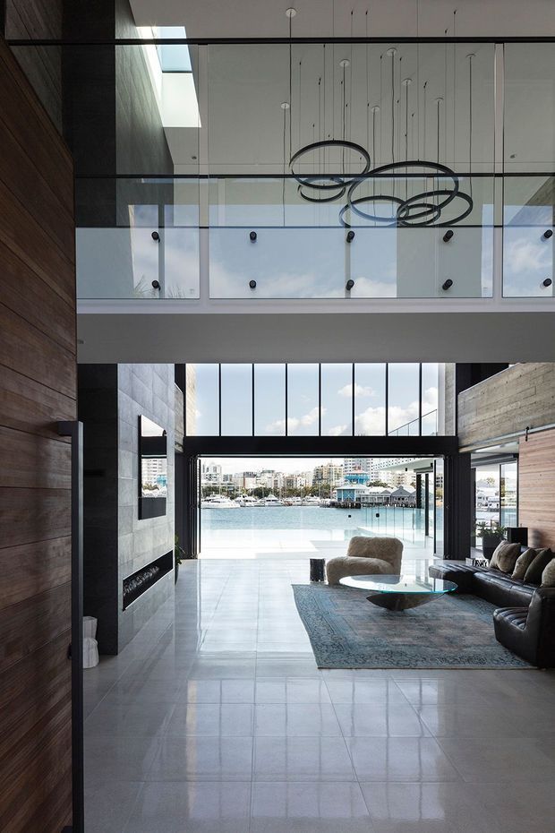A dramatic double-height living room with expansive floor-to-ceiling glazing opening toward a waterfront marina. The space features large-format polished light-grey floor tiles, dark timber wall panelling, exposed concrete or stone walls, a black-framed glazed mezzanine balustrade, and sculptural circular pendant lighting. A dark sectional sofa, armchair, coffee table, and patterned rug form the seating area, while the open glazing provides strong natural light and water views.