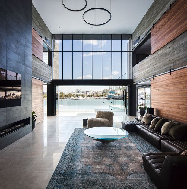A spacious double-height living room with dramatic floor-to-ceiling glazing opening toward a waterfront marina and urban skyline. The room features exposed concrete block and timber wall finishes, dark metal-framed glazing, polished light flooring, a large patterned rug, a dark leather sectional sofa, a round coffee table, a wall-mounted television and a linear fireplace. Circular pendant lights hang from the high ceiling, and an upper-level walkway overlooks the room.