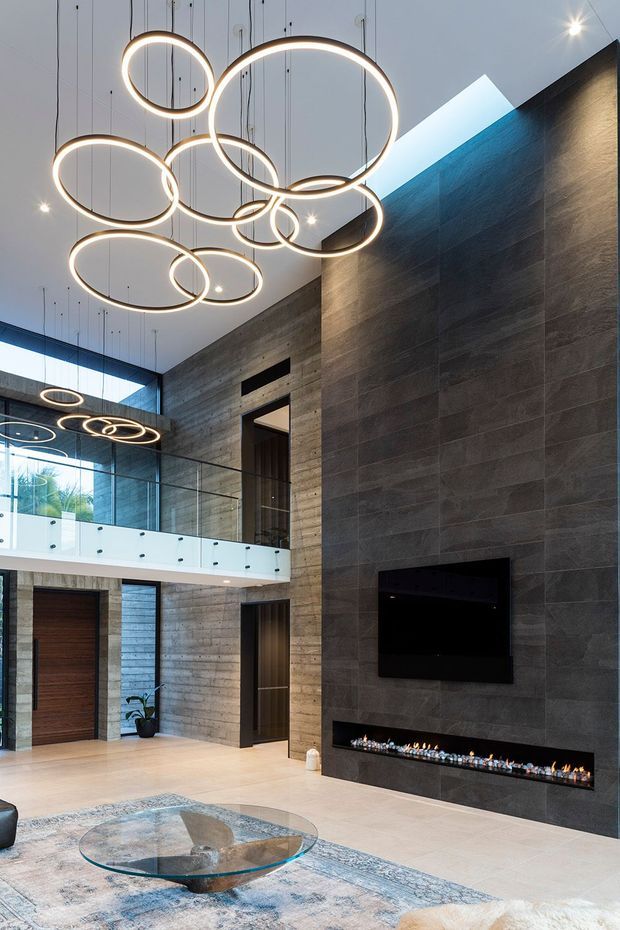 A dramatic double-height residential living space with a tall dark stone feature wall, linear gas fireplace and wall-mounted television. Sculptural circular pendant lights hang over a glass coffee table and rug, while a glazed upper-level balcony, exposed concrete-toned walls, timber doors and large windows contribute to the contemporary interior.
