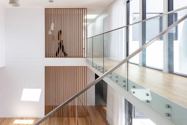 Bright contemporary multi-level interior focused on a stairway and landing, with extensive floor-to-ceiling glazing, pale timber flooring, clear glass balustrades, and diagonal metal handrails. Vertical timber screening forms a feature partition around the stair void, while daylight creates an airy, open atmosphere.