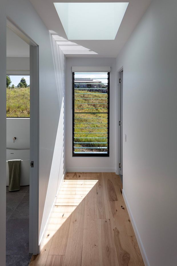 A bright, narrow residential hallway with pale painted walls, wide light timber floorboards, and a large skylight creating strong geometric sunlight and shadow. A louvred window at the end overlooks a grassy site and neighbouring building. An open doorway reveals part of a contemporary bathroom with dark tiled flooring, a freestanding or built-in white bathtub, and a towel. Simple white doors and trims complete the clean, minimal interior.