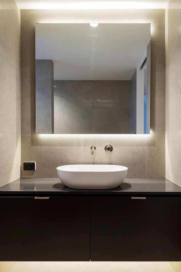 A compact contemporary bathroom vanity viewed straight on, featuring a white vessel basin on a dark stone benchtop, wall-mounted chrome tapware, a large backlit mirror, taupe-grey tiled walls, and dark built-in cabinetry below.