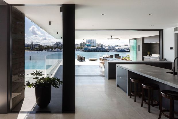 Open-plan contemporary kitchen and living area with a large dark island and integrated seating, opening through full-height glazed doors to a waterfront terrace and pool. The room features dark timber or wood-look cabinetry, a dark stone-look benchtop, pale large-format floor tiles, black framing, and extensive water-facing glazing. A lounge area and outdoor deck are visible beyond the kitchen.