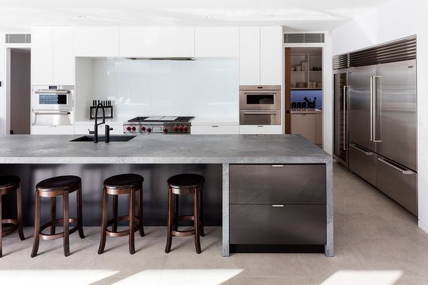 Bright, spacious contemporary kitchen dominated by a large island with a grey stone-look benchtop, dark integrated cabinetry, seating for several stools, stainless-steel appliances, and a pale glass splashback. White full-height cabinetry and a walk-in pantry or storage area are visible along the rear wall, with a large stainless-steel refrigerator at right.