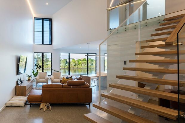 A bright, open-plan contemporary living area viewed from a floating timber staircase. The room features a large brown sectional sofa, armchairs, a timber media console, extensive glazing, and a dining or kitchen area beyond. The staircase has open timber treads, a glass balustrade, and a tall void with upper-level glazing. Polished speckled flooring, white walls, and linear integrated lighting create a clean, airy interior.