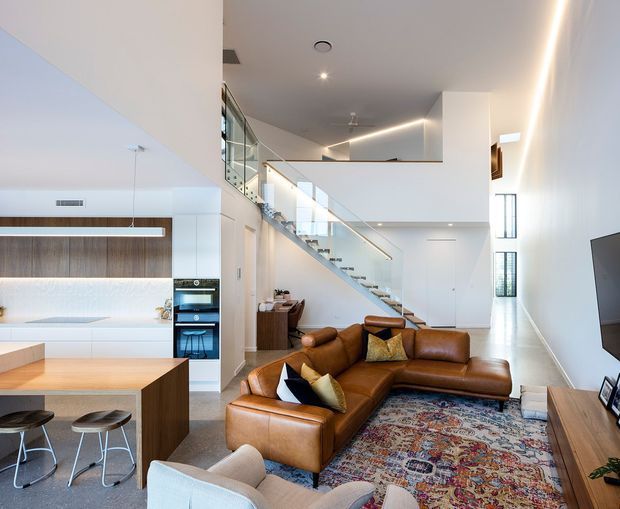 Bright, spacious open-plan contemporary interior with a kitchen and island in the foreground, a large brown leather sectional defining the living area, and a dramatic glass-balustraded staircase leading to an upper-level mezzanine. White walls, pale flooring, timber cabinetry, integrated appliances, recessed lighting and linear LED strip lighting create a clean modern setting, with a patterned rug adding warmth and colour.