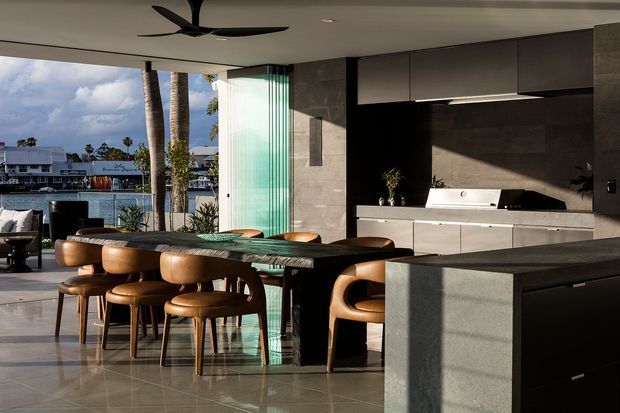 A refined open-plan kitchen and dining area with expansive glazing overlooking a waterfront setting. The room features dark, handleless kitchen cabinetry, a dark stone or concrete island, a long dark dining table with warm timber dining chairs, polished light flooring, and a sculptural black ceiling fan. A translucent fluted-glass screen separates the interior from the glazed opening, while strong natural light creates a high-contrast, sophisticated atmosphere.