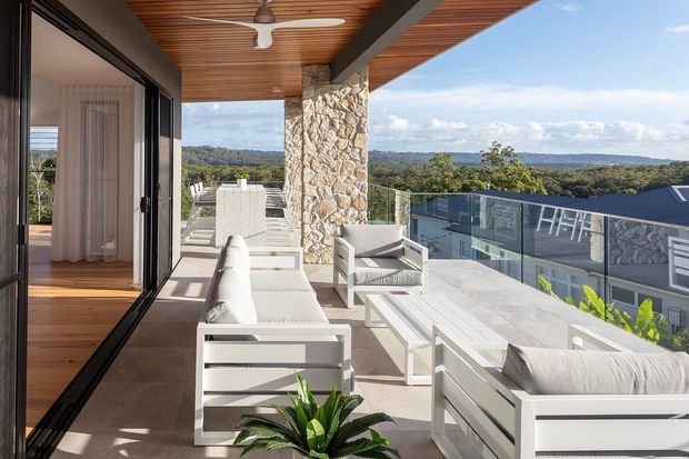 A spacious elevated outdoor terrace or balcony with white outdoor lounge furniture, a long dining table, glass balustrade, and a stone-clad structural column. The terrace has a warm timber-lined ceiling with a ceiling fan and opens directly to an interior through large black-framed sliding glass doors. A leafy landscape and neighbouring rooflines are visible beyond the railing.