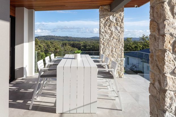 A covered outdoor dining terrace with a long white dining table and white chairs, framed by substantial rough natural-stone columns and a warm timber-lined ceiling. Pale large-format floor tiles, glass balustrading, and expansive views over a wooded landscape create a bright elevated entertaining area.