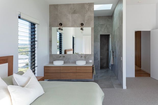 A bright contemporary bedroom with a partially visible bed in the foreground, opening directly onto a refined bathroom. The bathroom features a floating timber vanity with two white vessel basins, a large wall-to-wall mirror, grey stone-look tiled walls and floor, a walk-in shower, skylight, and louvred windows. Neutral finishes and strong natural light create a clean, calm atmosphere.