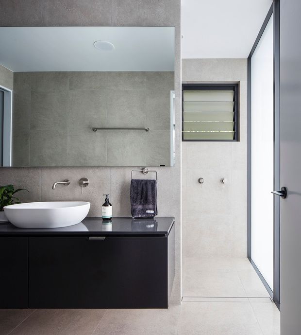 Contemporary bathroom featuring large-format grey-beige tiled walls and floor, a wall-mounted black vanity with a dark stone benchtop, white vessel basin, frameless mirror, wall-mounted tapware, and a separate shower area with a louvred window and glazed door. The space is clean-lined and brightly lit with strong black-and-neutral contrast.