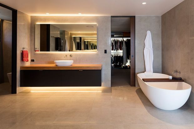 A luxurious contemporary bathroom with a freestanding white bathtub, wall-mounted dark vanity, vessel basin, large backlit mirror, and a walk-in wardrobe visible through an open doorway. The room features warm recessed and concealed lighting, large-format neutral stone-look surfaces, and minimalist integrated detailing.
