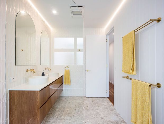 Bright contemporary bathroom with a long timber vanity, white vessel basin, brass wall-mounted tapware, arched frameless mirrors, white vertical wall tiles, pale stone-look floor tiles, and coordinated brass towel rails holding yellow towels. A frosted window and recessed ceiling lighting add to the clean, airy feel.