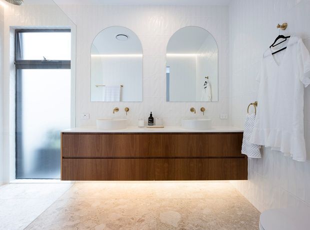 Bright contemporary bathroom with a double timber vanity, twin round-edged arched mirrors, two vessel basins, and a pale stone or marble floor. The walls feature subtle white textured vertical paneling, with a frosted-glass door or window at left and white robes hanging on brass hooks at right. The overall scheme is clean, warm, and refined.