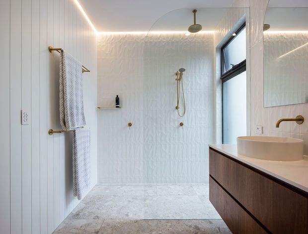 Bright contemporary bathroom with a walk-in shower, freestanding vessel basin, wall-mounted timber vanity, white textured wall panels, pale stone-look floor tiles, and brushed brass fixtures. A large mirror and window increase the sense of light and space, while concealed linear lighting adds a refined detail.