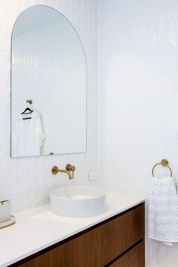 Bright, compact bathroom vanity featuring glossy white wall tiles, a white vessel basin on a white countertop, warm brass wall-mounted tapware and towel ring, a large arched mirror, and dark timber bathroom cabinetry.