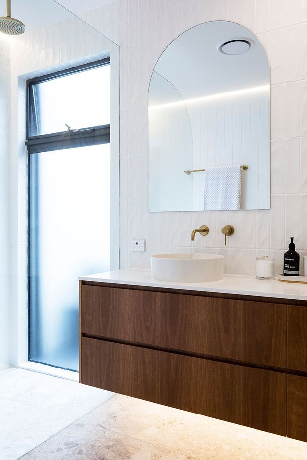 Bright contemporary bathroom featuring white tiled walls, a large arched mirror, a floating dark timber vanity with a white stone benchtop, round vessel basin, brushed brass wall-mounted tapware, and a frosted window. The space has clean lines and a refined, warm-modern finish.