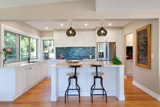 Bright, spacious white kitchen with a central island and two bar stools, white shaker-style cabinetry, stainless-steel refrigerator, patterned blue mosaic tile splashback, timber flooring, pendant lights, and large windows overlooking greenery. A chalkboard sign and pantry area are visible at the side.