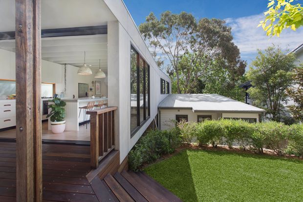 Exterior view of a contemporary multi-level home on a leafy sloped site, showing a dark timber deck and stair, white flat-roofed extension with extensive glazing, and an open-plan kitchen and dining area visible through the doors. A separate lower building with a corrugated metal roof sits among landscaped greenery.