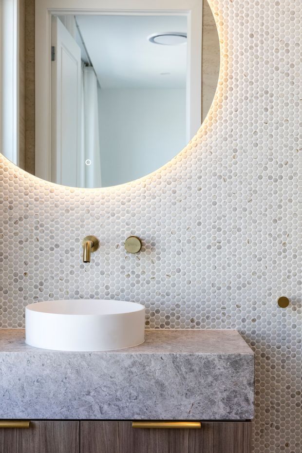 Contemporary bathroom vanity featuring a round white vessel basin on a grey natural-stone benchtop, dark timber cabinetry, brushed brass wall-mounted tapware, and a large round mirror with integrated backlighting. The walls are finished in small pale mosaic tiles, creating a refined, softly illuminated interior.