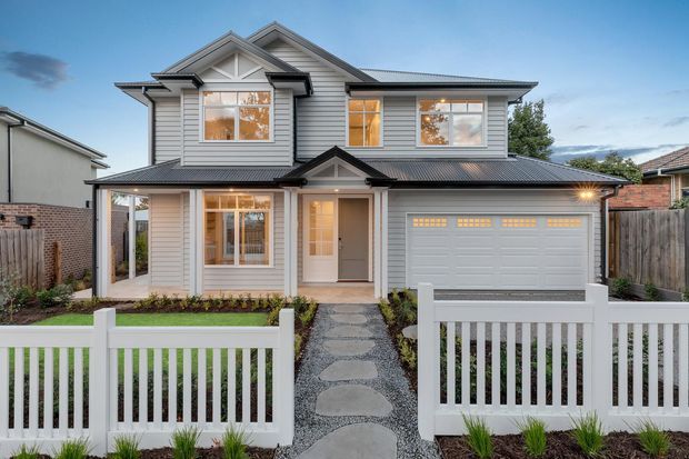 Front exterior view of a two-storey suburban residence with a symmetrical gabled roofline, light horizontal weatherboard cladding, dark grey roofing, white-framed windows, an attached double garage, covered entry porch, landscaped front garden, and white picket-style fencing. A paved stepping-stone path leads from the street-side fence to the entrance.