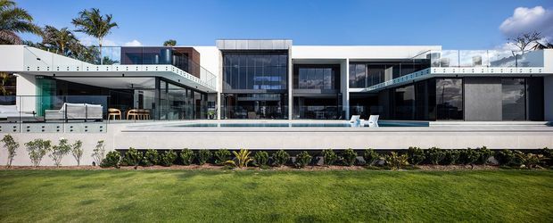 A wide exterior view of a large contemporary two-storey residence with strong horizontal lines, extensive floor-to-ceiling glazing, projecting cantilevered balconies, white rendered surfaces, dark window frames, and a long linear swimming pool in front. The landscaped lawn and tropical planting reinforce a resort-like setting.