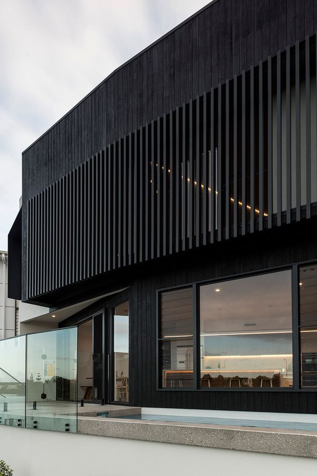 Exterior view of a contemporary multi-level building with dark vertical timber cladding and a prominent upper facade screened by closely spaced vertical fins. Large glazed openings reveal warm interior lighting, while a glass balustrade and paved terrace occupy the foreground. The composition emphasizes the bold black facade, projecting upper volume, and linear architectural detailing.