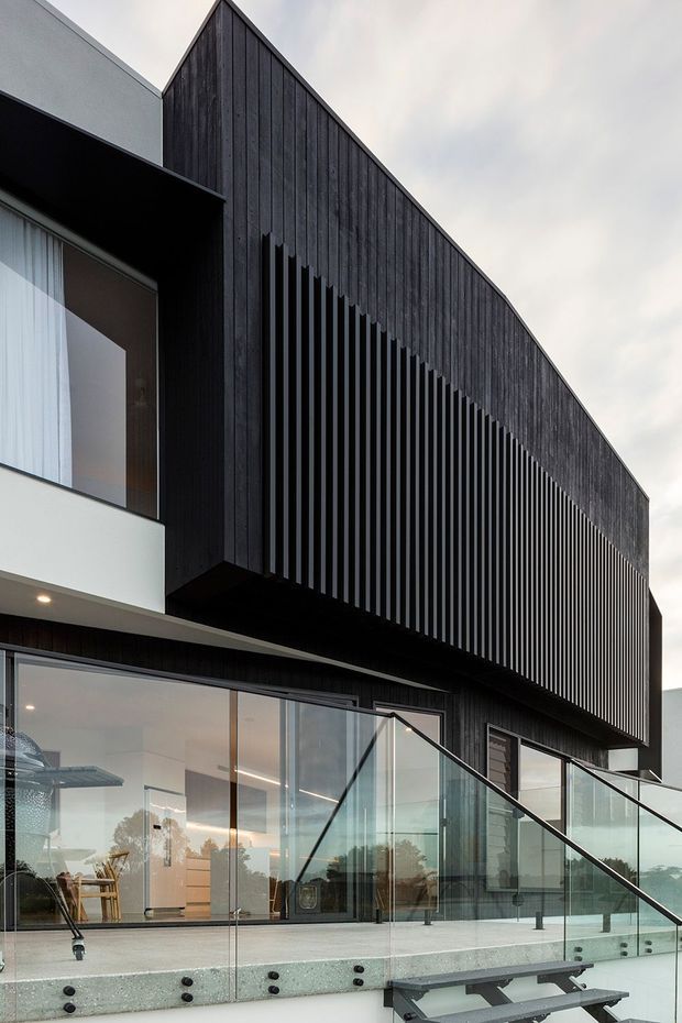 Exterior view of a contemporary multi-level residence with a dramatic steep gable-like dark timber-clad upper volume, vertical screening fins, large glazed openings, and a broad terrace with frameless glass balustrades and stairs. The lower level combines dark cladding, white rendered walls, and extensive floor-to-ceiling glazing. The image is photographed from below against an overcast sky.
