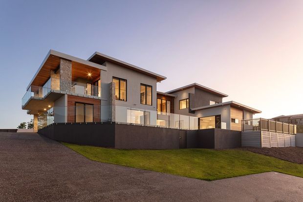 Exterior dusk view of a large contemporary multi-level residence with intersecting flat roof planes, projecting balconies, extensive glazing, warm timber soffits, stone cladding accents, and dark perimeter fencing. The home sits on a gently sloping landscaped site beside a paved street or driveway.
