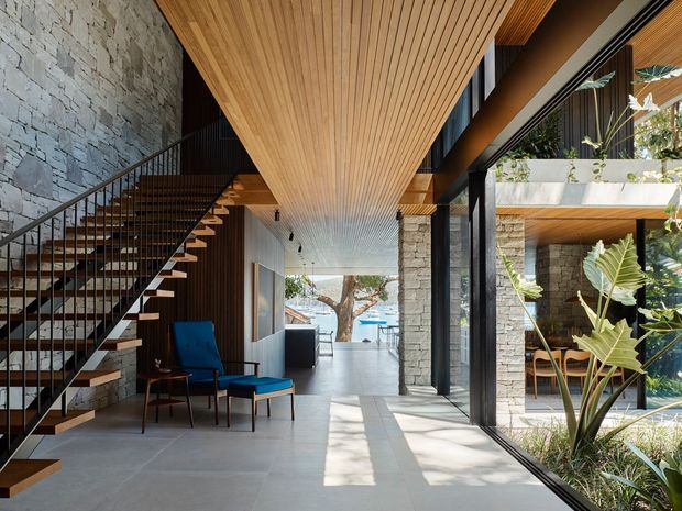 A refined contemporary interior centered on an open stairway and double-height circulation space. Floating timber stair treads with a slim black metal balustrade rise beside a textured natural-stone wall. Warm timber slatted ceilings and dark structural framing contrast with pale polished flooring and extensive floor-to-ceiling glazing. A lounge chair and ottoman sit beneath the stairs, while a planted courtyard and blue water feature are visible through the glazing.