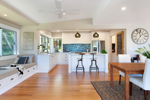 Bright open-plan interior combining a white kitchen with central island, living area, and dining space. The room features warm timber flooring, white cabinetry, a blue patterned tile splashback, pendant lights, large windows, and contemporary furnishings.