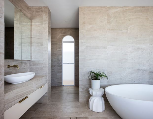 A refined contemporary bathroom featuring full-height beige natural-stone or stone-look wall and floor finishes, a freestanding white bathtub, floating vanity with integrated basin, large mirrored cabinetry, brushed brass tapware, and a narrow arched window. A sculptural white side table with a potted plant adds a decorative accent.