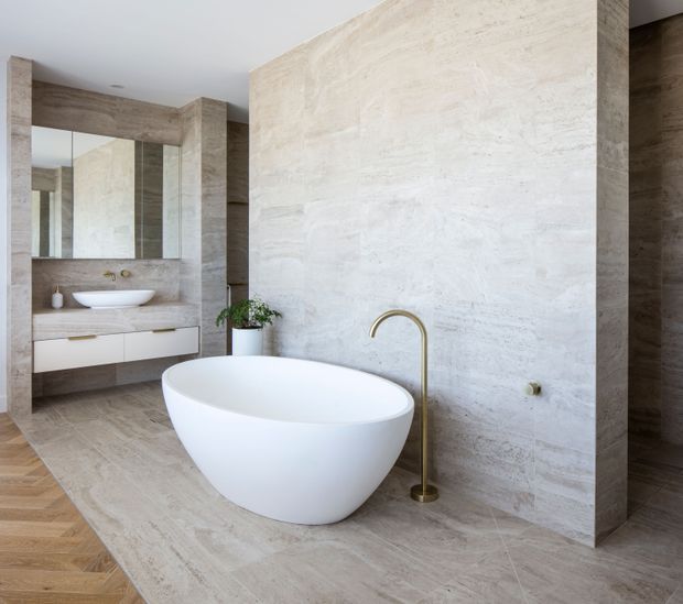 A bright, refined bathroom featuring a freestanding white oval bathtub, a brushed brass floor-mounted tub filler, pale stone-look wall and floor finishes, and a floating white vanity with a vessel basin. Large mirrors and timber-toned architectural framing add depth and warmth.