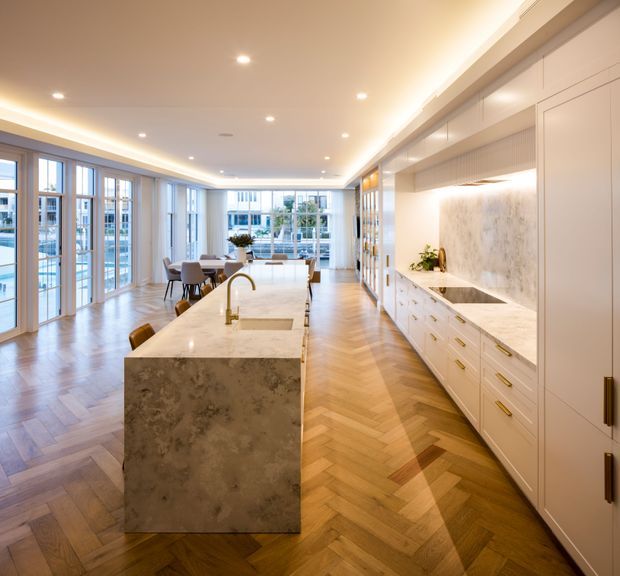 Bright, expansive open-plan kitchen and dining area with a large marble or stone island, extensive white built-in cabinetry, integrated appliances, and a patterned stone backsplash. Warm herringbone timber flooring, full-height glazed doors and windows, recessed ceiling lights, and concealed cove lighting create a refined contemporary interior.
