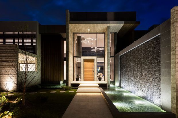 Night-time view of a contemporary residential facade and entry courtyard. A tall glazed entrance is framed by textured concrete or stone walls, with a warm timber front door, illuminated steps, a paved path, planted garden beds, and linear architectural lighting. The composition highlights the dramatic modern entry architecture and textured boundary wall.