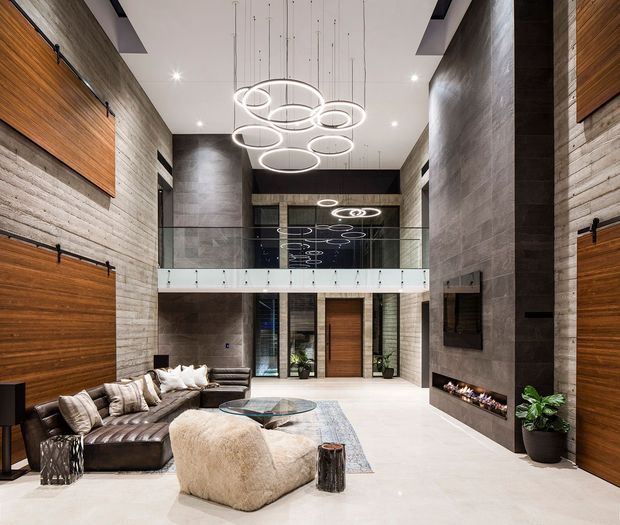 A dramatic double-height contemporary living room and entry hall with a mezzanine-level glass balustrade. The space features textured concrete-look walls, large horizontal timber wall panels and doors, a pale smooth floor, a suspended cluster of circular LED pendant lights, a large sectional sofa, glass coffee table, fireplace, and indoor plants.