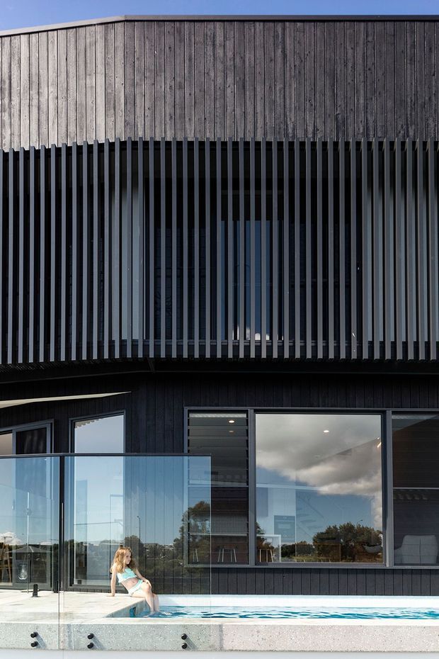 Exterior view of a contemporary multi-level residence with dark vertical cladding, a prominent timber-clad upper facade, and a deep vertical slatted screen enclosing the upper glazing. Large windows open toward a glass-balustraded pool terrace, with a swimming pool visible in the foreground. The composition highlights the building facade, screening, glazing, and outdoor pool area.
