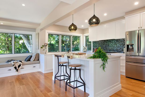 Bright, spacious kitchen with white shaker-style cabinetry, a large central island with seating, stainless-steel appliances, and a dark patterned mosaic tile backsplash. Warm timber flooring, abundant windows, recessed lighting, and decorative pendant lights create an inviting open-plan space. A built-in window seat is visible at the side.