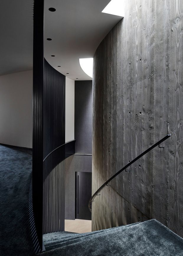 A dramatic, narrow interior stairway and landing with dark carpeted steps, a curved black metal handrail, ribbed dark wall lining, and a large textured concrete feature wall. A sculptural curved ceiling contains recessed downlights and bright skylight openings, creating a moody, highly architectural atmosphere.