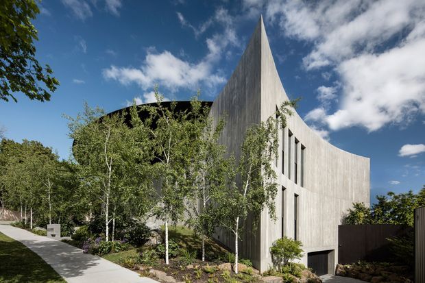 A striking sculptural concrete building set within landscaped grounds. The facade features tall, curved and sharply pointed forms, vertical textured concrete surfaces, narrow vertical windows, and a dark recessed entry or opening. A paved walkway and planted garden lead toward the building, with mature trees and a bright blue sky surrounding the structure.